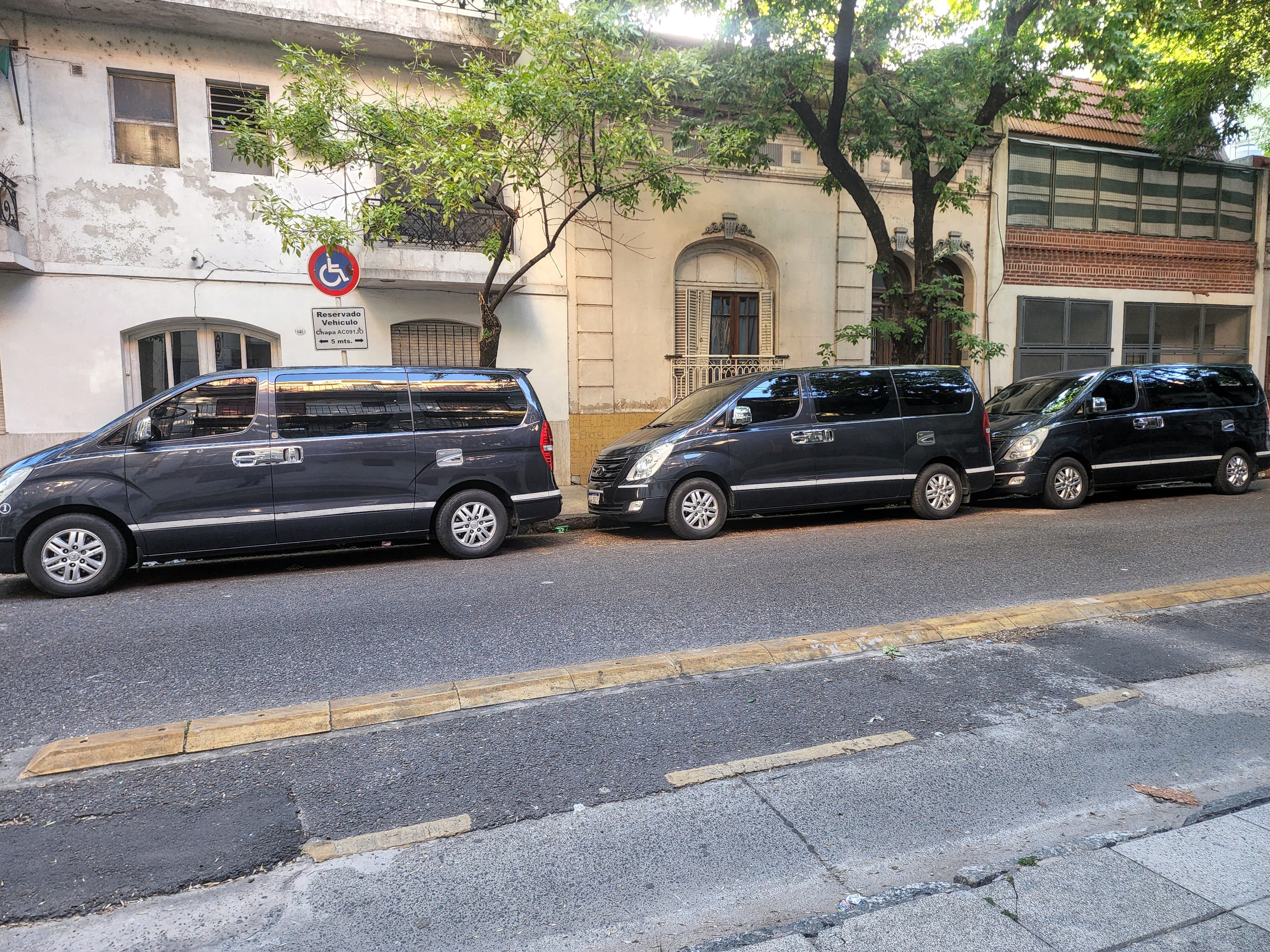 Tres furgonetas negras estacionadas en fila frente a edificios antiguos y árboles frondosos.